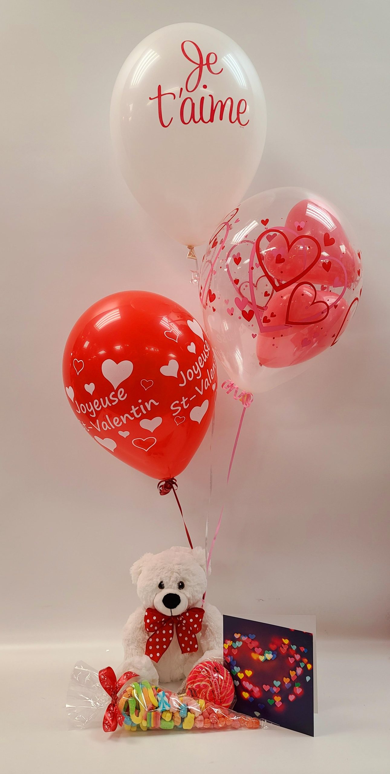 Trio de ballons dont 1 avec 2 coeurs dedans attachés au bras d'une peluche  avec 1 cône de bonbons, 1 suçon et une carte – ST16Les Folleries Fleuries